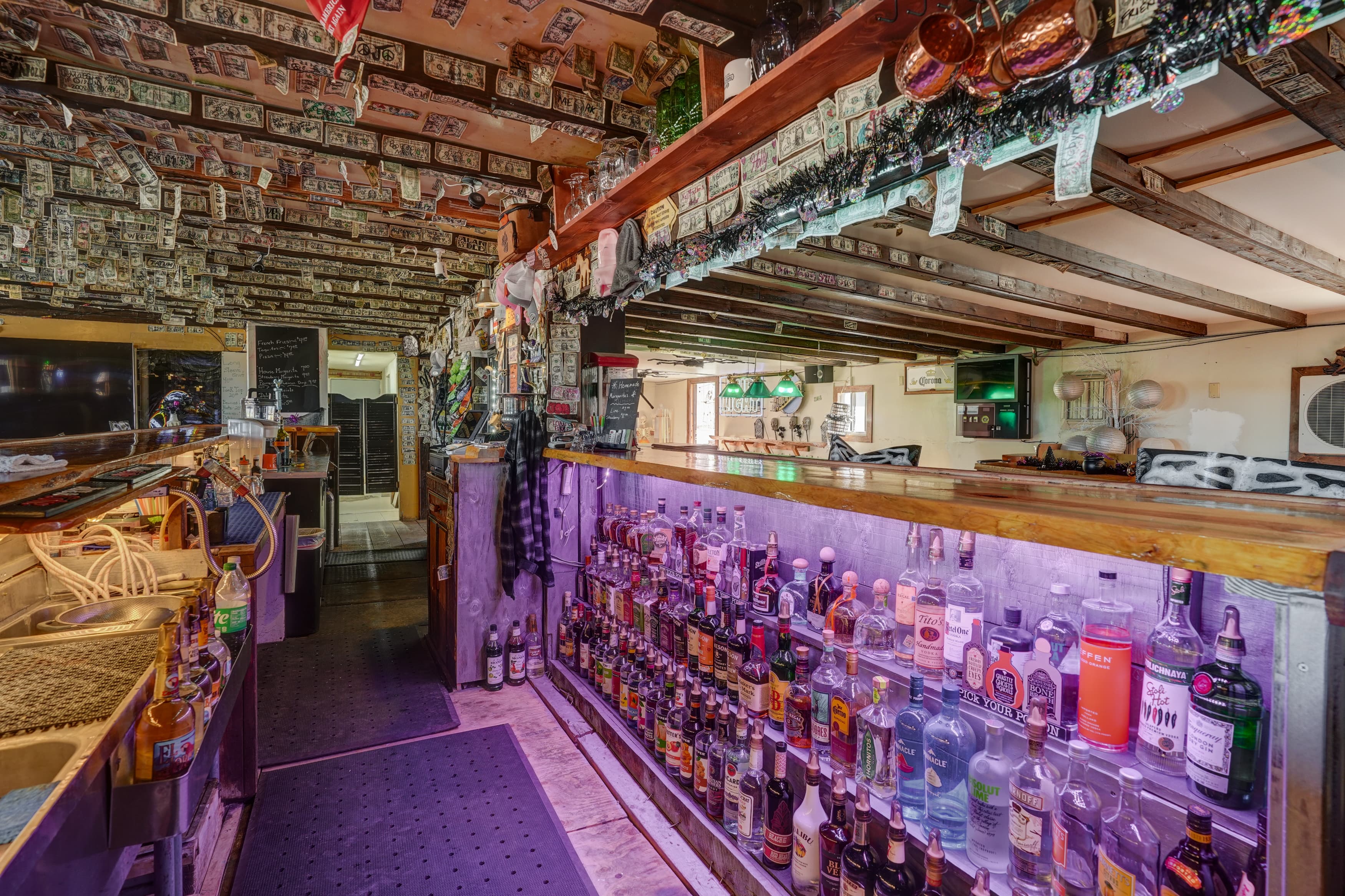 Empty wooden bar with purple-lit liquor bottles and currency notes covering the ceiling and walls.